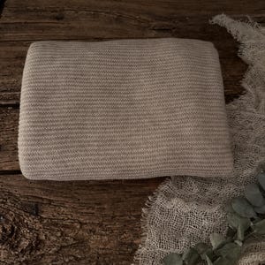 Peut inclure: Une couverture tricotée beige clair, pliée en rectangle. La couverture a une texture côtelée et est posée sur une surface en bois rustique. Un morceau de tissu beige transparent et de la verdure sont en arrière-plan.
