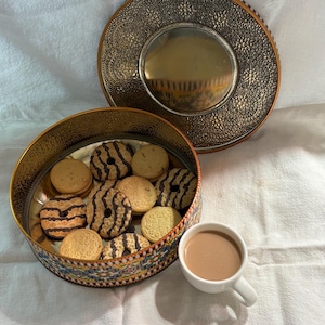 May include: An open, decorative tin filled with assorted cookies, some with chocolate stripes. A matching lid sits nearby. A small white cup of coffee sits next to the tin. The tin and lid have a gold and silver design.