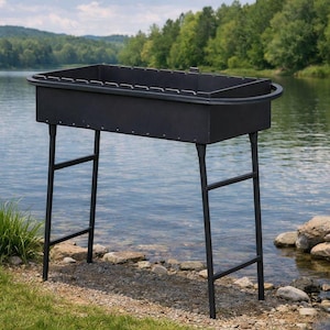 Op de afbeelding: Een zwarte metalen vuurplaats voor buiten met een rechthoekig bassin en een afgeronde handgreep. De vuurplaats wordt ondersteund door vier schuine poten. Het staat op een grasveld bij een meer, met bomen op de achtergrond.