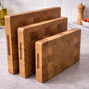 May include: Three wooden cutting boards of varying sizes, arranged vertically. The boards are made of a light brown wood with a block pattern and have a handle cutout. The boards are on a gray countertop.