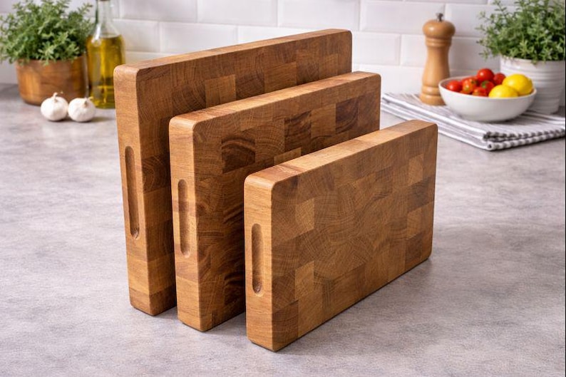 May include: Three wooden cutting boards of varying sizes, arranged vertically. The boards are made of a light brown wood with a block pattern and have a handle cutout. The boards are on a gray countertop.