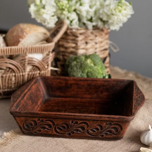 Può includere: Una pirofila rettangolare in ceramica marrone-rossastra con motivi floreali decorativi sui bordi. Il piatto è appoggiato su un tessuto beige, con un cestino di pane, un cesto di fiori e broccoli sullo sfondo.