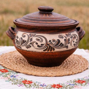 Op de afbeelding: Een handgemaakte, bruine keramische pot met een deksel, met een crèmekleurige band met een gedetailleerd bloemmotief. De pot heeft twee handvatten en staat op een geweven mat en geborduurde doek, wat een rustieke uitstraling suggereert.