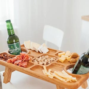 Könnte beinhalten: Ein Holzserviertablett mit mehreren Fächern, das verschiedene Snacks wie Käse, Nüsse und Wurstwaren enthält. Zwei Flaschen Bier und Wein stehen auf dem Tablett. Das Tablett hat klappbare Beine.