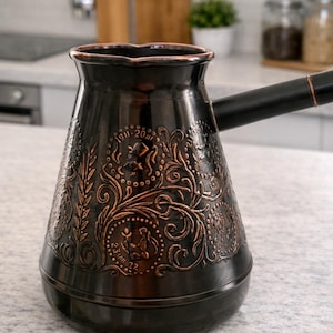 Könnte beinhalten: Eine dunkelbraune türkische Kaffeekanne mit einem verzierten kupferfarbenen Blumendesign. Die Kanne hat einen langen schwarzen Griff und steht auf einer hellen Arbeitsplatte. Der Text "1911-20..." und "23/VII/22." ist sichtbar.