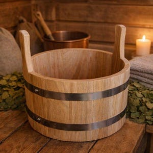 Könnte beinhalten: Ein hölzerner Saunaeimer mit zwei Griffen und Metallbändern, auf einer Holzoberfläche. Die Szene umfasst einen Saunahut, Handtücher, einen Kupfertopf und eine brennende Kerze, die eine warme, spa-ähnliche Atmosphäre schafft.