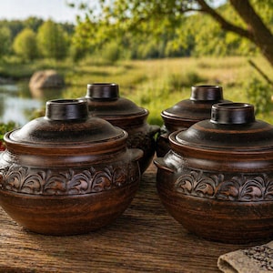 May include: Four brown ceramic cooking pots with lids, featuring a decorative floral pattern around the base. The pots are arranged on a wooden surface, with a blurred outdoor background of trees and a lake.