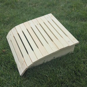 Può includere: Un poggiapiedi in legno chiaro con un design curvo. La parte superiore è realizzata con doghe di legno parallele e i lati sono pieni. Il poggiapiedi è posto su erba verde.