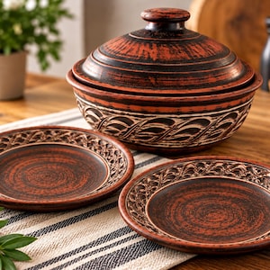 Op de afbeelding: Een set van donker roodbruin aardewerk servies bestaande uit een kom met deksel en twee bijpassende borden. Elk stuk is voorzien van een gestructureerd, uitgesneden bladerpatroon langs de rand en een rustieke, gevlekte afwerking.