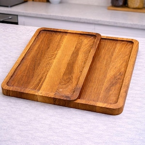 Può includere: Due vassoi rettangolari in legno con una calda venatura naturale. I vassoi hanno un bordo rialzato e sono impilati su una superficie di colore chiaro. Gli utensili da cucina sono ideali per servire cibo o esporre oggetti.