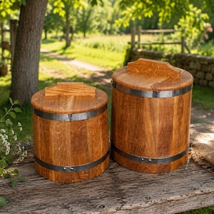 Könnte beinhalten: Zwei zylindrische Holzbehälter mit Deckeln, die eine natürliche braune Holzmaserung und dunkle Metallbänder oben und unten aufweisen. Jeder Deckel hat einen kleinen rechteckigen Griff. Die Behälter stehen nebeneinander auf einer Oberfläche.