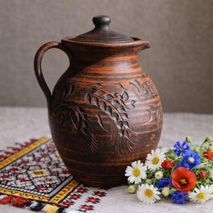Può includere: Una brocca rustica in ceramica marrone con coperchio e manico, con disegni di spighe di grano intagliate. La brocca è posta su un tessuto fantasia accanto a un piccolo bouquet di fiori di campo, tra cui margherite e papaveri rossi.
