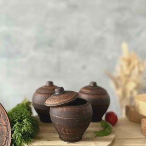 Peut inclure: Trois pots en céramique brun foncé avec couvercles, dont un partiellement ouvert, sont posés sur une planche en bois. Les pots présentent un design rustique et texturé. De l'aneth frais, une tomate et un poivron sont également visibles.