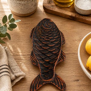 Könnte beinhalten: Eine Löffelablage aus Gusseisen in Fischform mit mattschwarzer Oberfläche und kupferfarbenen Akzenten an den erhabenen Schuppen- und Flossendetails. Das Objekt weist eine strukturierte Oberfläche mit ausgeprägtem Kopf, Körper und Schwanz auf.