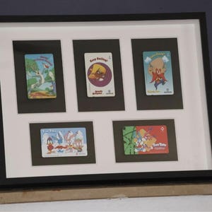 Könnte beinhalten: Ein schwarzer rechteckiger Rahmen mit fünf Looney Tunes-Sammelkarten in einer Passepartout-Anordnung. Die Karten zeigen verschiedene Zeichentrickfiguren mit Texten wie "Keep Smiling!", "Speedy Gonzales" und "Tiny Toons Adventures".