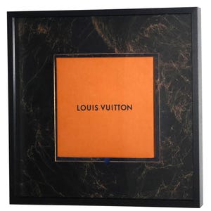 Könnte beinhalten: Ein quadratischer schwarzer Rahmen mit einem schwarzen und kupferfarbenen Marmormuster-Hintergrund. Zentriert im Rahmen befindet sich eine leuchtend orangefarbene quadratische Platte mit dem schwarzen Text LOUIS VUITTON und einer kleinen blauen Lasche.