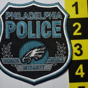 May include: A shield-shaped embroidered patch featuring the text PHILADELPHIA POLICE in light blue, an eagle graphic, and the words HONOR, SERVICE, and INTEGRITY. A yellow ruler is visible, showing the patch is approximately 10 cm in height.