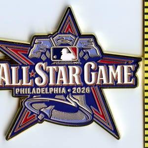 May include: A star-shaped pin with a gold border, featuring the text "ALL STAR GAME PHILADELPHIA 2026." The pin has a blue and red color scheme with the MLB logo. The pin is next to a yellow ruler.
