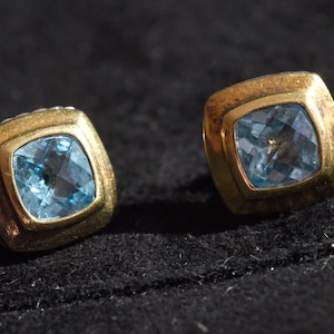 May include: Two square-shaped stud earrings with gold-colored frames and light blue gemstones. The earrings are set against a dark background, highlighting their design and color.