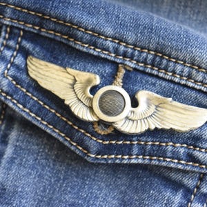 May include: A silver-toned metal pin shaped like a pair of wings with a central circular emblem featuring horizontal lines, attached to the flap of a blue denim pocket with visible orange stitching along the edges of the fabric.