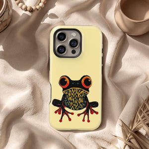 May include: A yellow phone case featuring a stylized frog illustration. The frog has large orange eyes, a black body with yellow and black details, and red feet. The case has a glossy finish and a camera cutout.