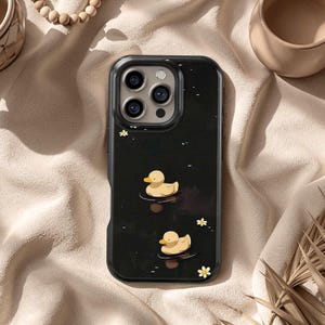 Puede incluir: Funda de teléfono negra con un diseño caprichoso de dos patitos de goma amarillos flotando en el agua. La funda tiene un acabado brillante y está adornada con pequeños acentos de flores blancas, creando una estética lúdica.