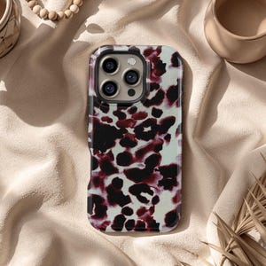 May include: A smartphone case with a white background and a pattern of black and burgundy spots. The case has a black border and a camera cutout. The case is lying on a beige surface.