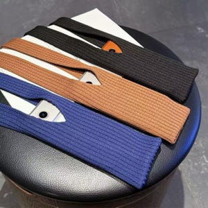 May include: Four knitted phone holders in black, brown, tan, and blue. Each holder has a pocket for a phone and a small leather accent. The holders are arranged on a black, round surface.