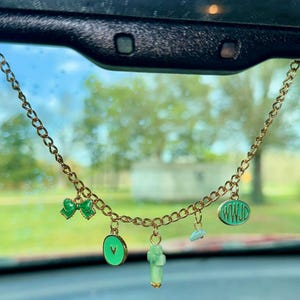 May include: A gold-colored chain with multiple charms, including a green bow, a green circle with an arrow, a green cross, a blue stone, and a green oval with the letters "WWJD". The charms are hanging in front of a car's windshield.