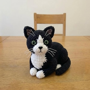 Könnte beinhalten: Eine handgefertigte Häkelkatze in Schwarz und Weiß, mit grünen Augen und weißen Schnurrhaaren. Die Katze liegt auf einer Holzoberfläche. Die Katze hat einen weißen Fleck im Gesicht und weiße Pfoten.
