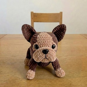 Puede incluir: Un juguete hecho a mano de ganchillo que representa un perro bulldog francés, en marrón y beige. El peluche tiene orejas grandes, una nariz negra y ojos grandes. El perro está tumbado sobre su vientre.