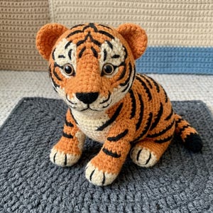 May include: A handmade crocheted tiger cub, featuring orange and black stripes, cream-coloured details, and large eyes. The plush toy is positioned on a grey, textured surface, highlighting the intricate crochet work.