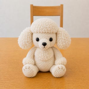 May include: A cream-colored crocheted poodle stuffed animal featuring a textured curly yarn head and ears, smooth yarn body, black plastic eyes, and a small black nose. The toy is seated in an upright position with visible paw pad details.