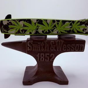 May include: A folding knife with a green and black cannabis leaf design, resting on a brown anvil-shaped stand. The stand is embossed with "Smith & Wesson 1852". The knife's handle features green cannabis leaves and skull designs.