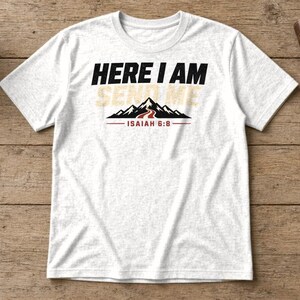 Pu&ograve; includere: Una t-shirt bianca a maniche corte con il testo nero HERE I AM, il testo beige SEND ME e un grafico di montagne nere con un sentiero rosso. Sotto il grafico della montagna c'&egrave; il testo rosso ISAIAH 6:8 con una linea rossa su ogni lato.
