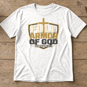 May include: A white short-sleeve t-shirt featuring a graphic design of a shield with a cross at the top. The text reads FULL ARMOR OF GOD and EPHESIANS 6:11 in gold and dark gray lettering centered on the front of the garment.