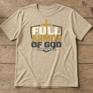 May include: A heathered beige short-sleeve t-shirt featuring a graphic of a shield with a cross and the text "FULL ARMOR OF GOD EPHESIANS 6:11" in gold and dark gray lettering centered on the chest.
