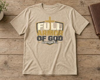 May include: A heathered beige short-sleeve t-shirt featuring a graphic of a shield with a cross and the text "FULL ARMOR OF GOD EPHESIANS 6:11" in gold and dark gray lettering centered on the chest.