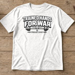 Puede incluir: Una camiseta blanca de manga corta con texto negro que dice TRAINED HANDS FOR WAR, un gr&aacute;fico de una barra de pesas con una cruz en el centro y el texto PSALM 144:1 impreso debajo del gr&aacute;fico de la barra de pesas.