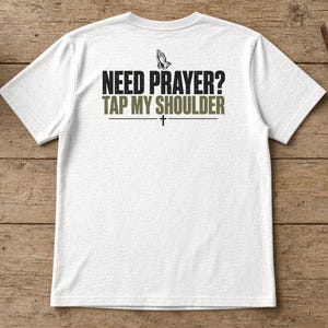 May include: A white short-sleeved t-shirt featuring a graphic on the back with the text "NEED PRAYER? TAP MY SHOULDER" in black and olive green lettering, accompanied by an icon of praying hands and a small cross symbol.