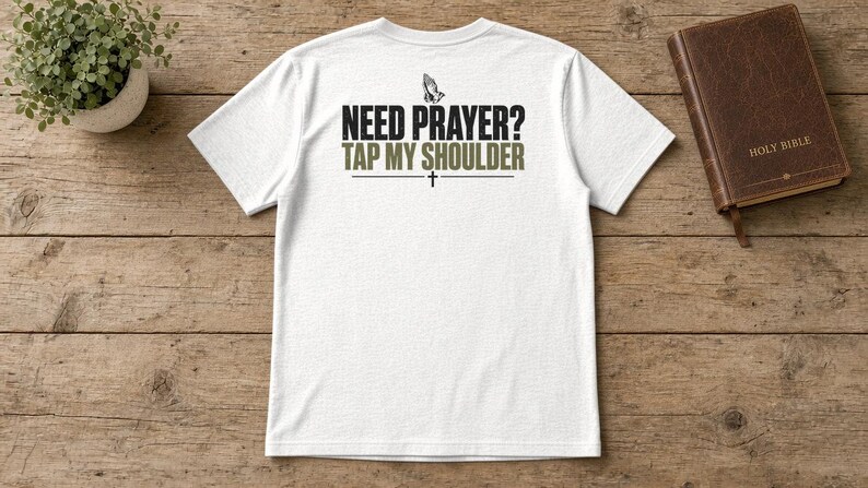 May include: A white short-sleeved t-shirt featuring a graphic on the back with the text "NEED PRAYER? TAP MY SHOULDER" in black and olive green lettering, accompanied by an icon of praying hands and a small cross symbol.