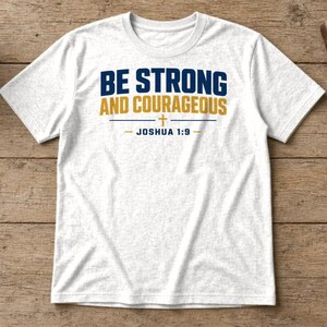 May include: A white short-sleeve t-shirt featuring the dark blue and gold text BE STRONG AND COURAGEOUS with a small cross symbol and the text JOSHUA 1:9 printed in dark blue on the front centre of the garment.