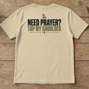 May include: A beige short-sleeve t-shirt featuring a graphic on the back with the text "NEED PRAYER? TAP MY SHOULDER" in black and olive green lettering, accompanied by an illustration of praying hands and a small cross symbol.
