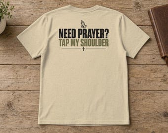 May include: A beige short-sleeve t-shirt featuring a graphic on the back with the text "NEED PRAYER? TAP MY SHOULDER" in black and olive green lettering, accompanied by an illustration of praying hands and a small cross symbol.