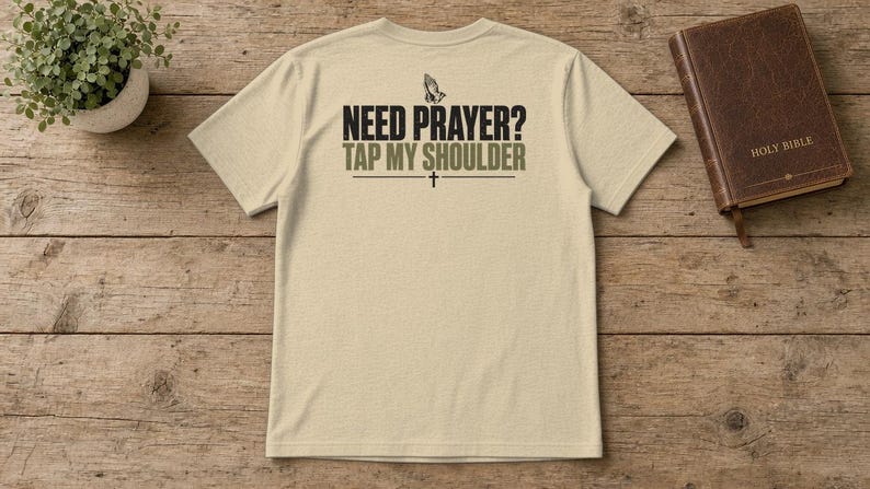 May include: A beige short-sleeve t-shirt featuring a graphic on the back with the text "NEED PRAYER? TAP MY SHOULDER" in black and olive green lettering, accompanied by an illustration of praying hands and a small cross symbol.