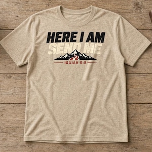 Pu&ograve; includere: Una t-shirt a maniche corte color beige m&eacute;lange con il testo in bianco e nero "HERE I AM SEND ME" sopra un grafico di montagne nere con un sentiero rosso, e il testo "ISAIAH 6:8" sottolineato in rosso sotto il grafico.