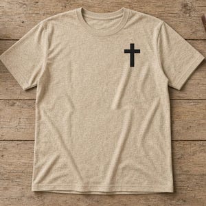 May include: A heathered beige short-sleeve t-shirt featuring a simple black cross graphic printed on the upper left chest area. The shirt has a crew neckline and a standard fit, showcasing a neutral tone and a clean, minimalist design.