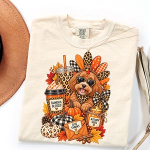 May include: A cream-colored t-shirt featuring a graphic of a dog wearing heart-shaped sunglasses and a turkey-feather headdress. The design includes a coffee cup labeled Thankful Blessed, a candle labeled Autumn Vibes, and an envelope labeled Give Thanks.