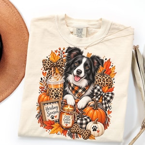 May include: A cream-colored t-shirt featuring a graphic of a black and white Border Collie wearing a plaid bandana, surrounded by pumpkins, autumn leaves, a leopard print bow, a coffee cup, and a candle with text reading Herding Season.