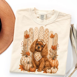May include: A cream-colored t-shirt featuring a graphic of a brown spaniel dog wearing a patterned bandana and hair bow. The dog sits before a wooden fence surrounded by orange autumn leaves and several patterned pumpkins in shades of orange.
